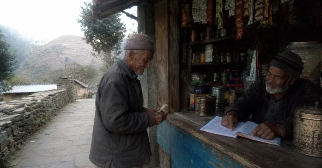 पहाड़ी गाँव की राशन दुकान में उधार की कॉपी देखते दुकानदार और हिसाब चुकता करते बुजुर्ग ग्रामीण का दृश्य