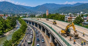 देहरादून में निर्माणाधीन 12 Km लंबा आशारोड़ी-झाझरा बायपास कॉरिडोर, जहां चार-लेन सड़क का कार्य जारी है और आसपास हरियाली दिखाई दे रही है