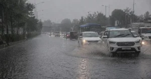 बारिश के बाद सड़क पर जमा पानी के बीच सावधानी से गुजरते वाहन और सक्रिय मानसूनी मौसम का दृश्य।