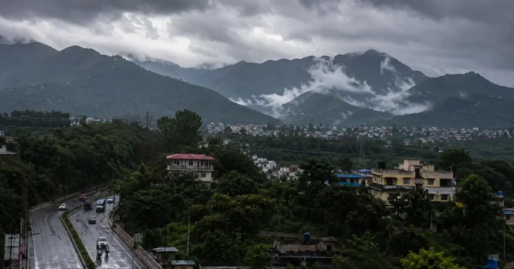 उत्तराखंड के पहाड़ी शहर में काले बादलों, धुंध और बारिश के बीच सड़क पर चलता ट्रैफिक