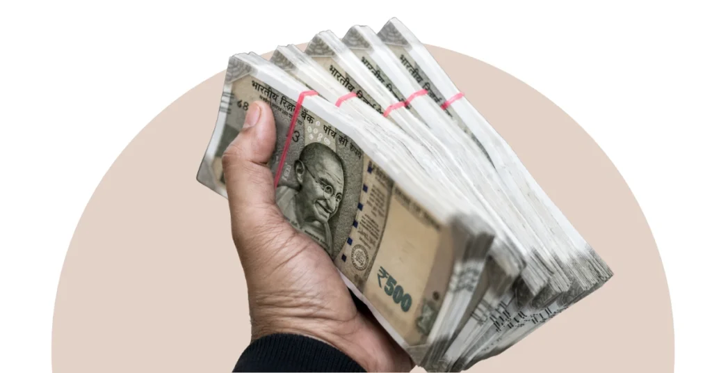 A hand holding multiple bundles of Indian currency notes used as a representative visual for an alleged financial irregularity investigation.