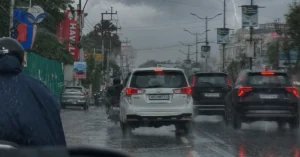 Vehicles and two-wheelers move through heavy rain on a city road in Uttarakhand as dark clouds and lightning reduce visibility.