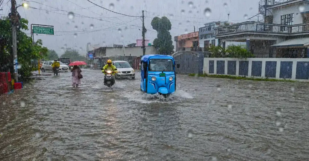 A flooded urban street in Uttarakhand during heavy monsoon rain, with an auto-rickshaw, motorcycles and pedestrians moving cautiously through waterlogged roads.