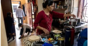 A woman preparing food in a home kitchen while other family members continue their morning routine in the background.