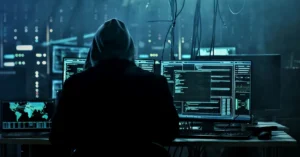 A hooded individual working at multiple computer screens, representing cyber fraud and online financial scams.