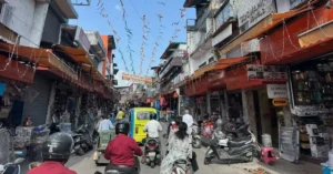 Paltan Bazaar in Dehradun showing scooters, a three-wheeler, pedestrians and shops along a busy market street.