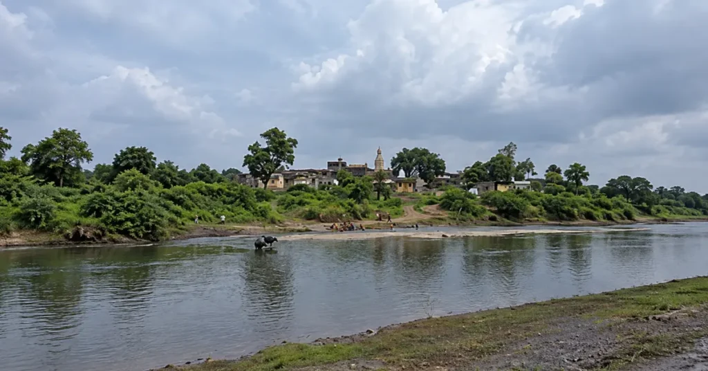 View of the Chandrabhaga River in Rishikesh, Uttarakhand, showing a calm river channel, green riverbanks, scattered buildings and an overcast sky.