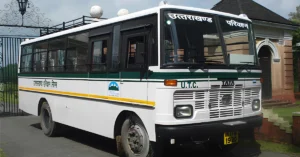 White Uttarakhand Transport Corporation (UTC) bus with dark green window panels and a yellow side stripe parked outside a gated building.